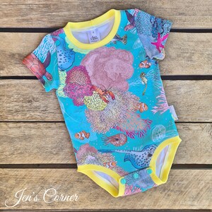 May include: A turquoise baby onesie with a yellow trim and snaps. The onesie features a colorful print of sea life, including coral, fish, and a crab.