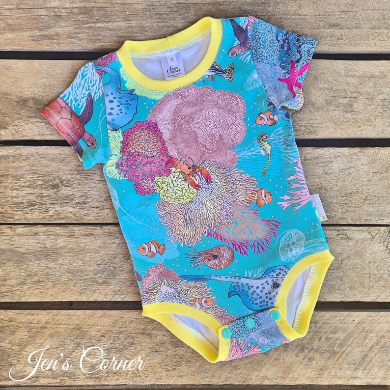 May include: A turquoise baby onesie with a yellow trim and snaps. The onesie features a colorful print of sea life, including coral, fish, and a crab.