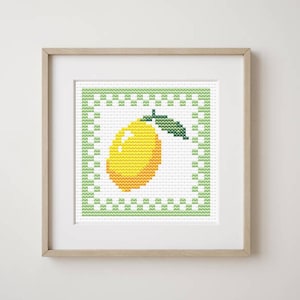 May include: A framed cross-stitch artwork featuring a yellow lemon with a green leaf. The lemon is centered within a white background, framed by a green border with a repeating pattern. The artwork is displayed in a light-colored wooden frame.