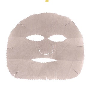 Puede incluir: Una mascarilla facial de tela color beige claro con aberturas para los ojos, la nariz y la boca. Sobre la mascarilla hay un pequeño icono de una bañera con una flor de loto y puntos amarillos. Debajo, el texto dice ONLY GOOD BAIN PARIS.
