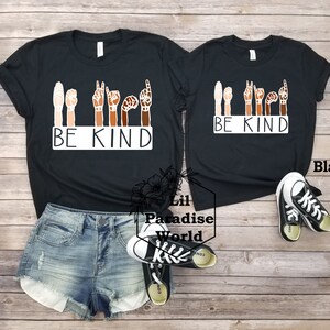 May include: Two black t-shirts with the phrase "BE KIND" in white lettering. The phrase is signed in American Sign Language with five hands of different skin tones. The shirts are on a wooden surface with a pair of black and white sneakers.