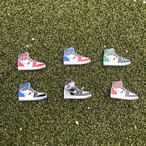 May include: Six enamel pins shaped like high-top sneakers arranged in two rows. The pins feature various color combinations including red and white, blue and white, green and white, blue and black, grey and black, and pink and white with black details.