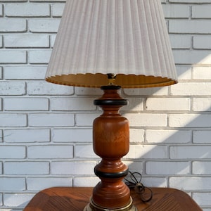 May include: A table lamp featuring a pleated off-white fabric lampshade and a turned wooden base with natural wood grain, accented by two black rings and a circular brass-toned metal pedestal at the bottom, including a black electrical cord.