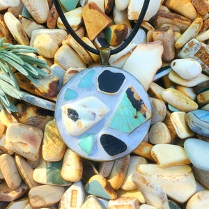 May include: A round pendant necklace with a black cord. The pendant features a mosaic design with various shapes of turquoise, black, and white stones set in a light blue background. The pendant is displayed on a bed of colorful pebbles.