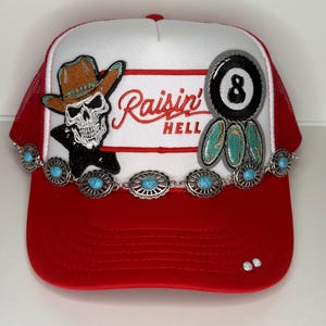 Könnte beinhalten: Rote und weiße Trucker-Mütze mit einem Totenkopf mit Cowboyhut und dem Text "Raisin' Hell". Mit einer 8-Ball und türkisfarbenen Akzenten. Die Mütze hat eine rote Mesh-Rückseite und einen gebogenen Schirm.