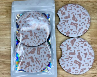 May include: Two white round car coasters with a pink cowboy boot, hat, and star pattern. The coasters are in a clear plastic bag with a holographic iridescent background.