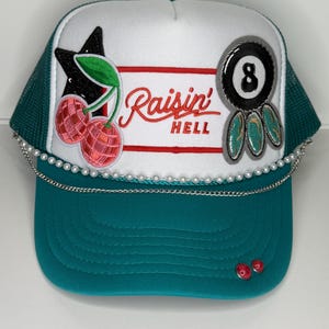 Puede incluir: Gorra de camionero turquesa y blanca con una cadena de perlas y varios parches. Los parches incluyen una estrella negra, cerezas, una bola ocho y las palabras "Raisin' Hell" en rojo.