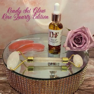 May include: Ready Set Glow Rose Quartz Edition beauty set with a rose quartz roller, eye masks, and a bottle of 24k Rose Gold Beauty Oil. The set is displayed on a gold mirrored cake stand.