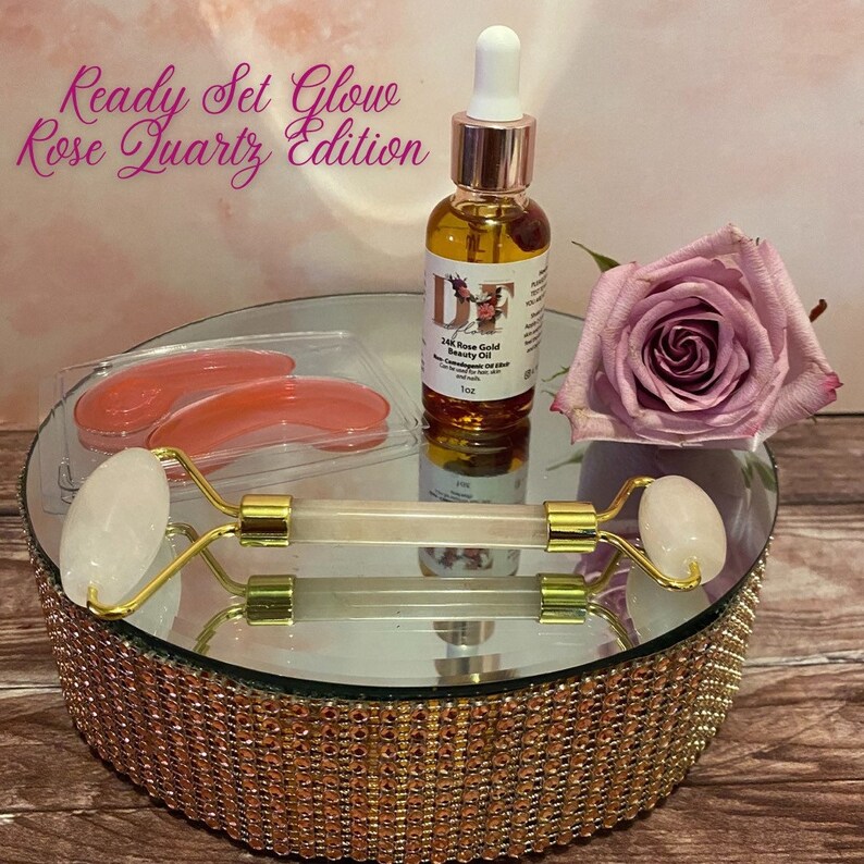 May include: Ready Set Glow Rose Quartz Edition beauty set with a rose quartz roller, eye masks, and a bottle of 24k Rose Gold Beauty Oil. The set is displayed on a gold mirrored cake stand.
