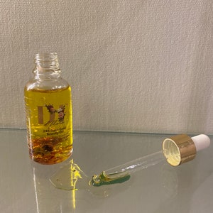 May include: A clear glass bottle of d'flora 24K Rose Gold Beauty Oil with a gold-toned dropper cap. The bottle contains yellow oil with visible gold flakes and floral petals. A separate glass dropper rests nearby with a drop of oil on the surface.