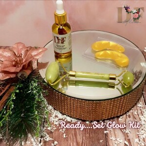 May include: A gold and green beauty set on a mirrored tray with a gold trim. The set includes a jade roller, a bottle of 24k Rose Gold Beauty Oil, and a pair of gold under-eye patches. The tray is on a wooden surface with white confetti. The text "Ready...Set Glow Kit" is on the bottom of the image.