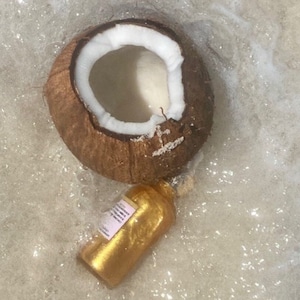 May include: A halved brown coconut with a white interior and a small gold-colored bottle with a rectangular label, both partially submerged in a bubbly, light-colored liquid. The coconut shell has a textured, fibrous exterior surface.