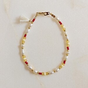 May include: A delicate bracelet with white, yellow, and red beads, accented with a small white tassel.