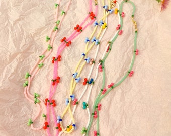 May include: Five beaded necklaces with different colored beads and charms. The necklaces are made with small, colorful beads and feature small charms in the shape of cherries. The necklaces are laid out on a pink surface.