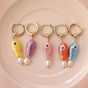 May include: Five pairs of gold hoop earrings with a pearl and a ceramic fish charm. The fish charms are in different colours: yellow, purple, pink, orange, and blue.