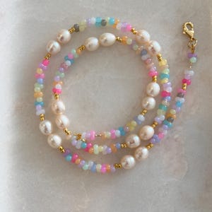 Könnte beinhalten: Eine Halskette bestehend aus einer Reihe weißer Süßwasserperlen, die mit kleinen, bunten Glasperlen in Rosa, Blau, Gelb und Violett durchsetzt sind. Das Design enthält winzige goldfarbene Zwischenperlen und einen goldfarbenen Karabinerverschluss.