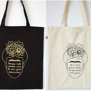 Könnte beinhalten: Zwei Einkaufstaschen mit einem Schwarz-Weiß-Design von Frida Kahlo mit Blumen im Haar. Die schwarze Tasche hat goldenen Text, der "Porque todo cambio nace de una gran destrucción" lautet. Die weiße Tasche hat schwarzen Text, der "Ni tú eres para tanto, Ni yo para tan poco" lautet.