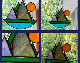 May include: A stained glass suncatcher depicting a mountain range with a blue lake and a bright orange sun. The suncatcher is hanging from a chain.