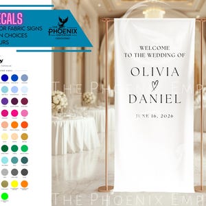 May include: A white wedding banner with the text "WELCOME TO THE WEDDING OF OLIVIA & DANIEL JUNE 16, 2026". The banner is displayed on a gold stand. The image also shows a colour chart and text that reads "IRON ON DECALS FOR FABRIC SIGNS".