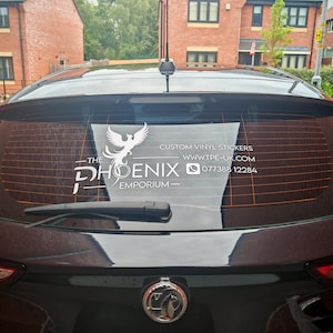 May include: A black car's rear window displays a custom vinyl sticker. The white sticker features a phoenix logo and the text "The Phoenix Emporium", along with contact details for custom vinyl stickers.
