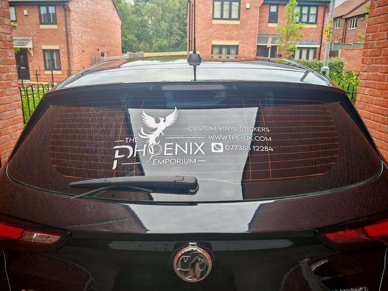 Pode incluir: Vista traseira de um carro preto com um autocolante de vinil personalizado no vidro traseiro. O autocolante branco apresenta um log&oacute;tipo de f&eacute;nix e o texto "The Phoenix Emporium" e informa&ccedil;&otilde;es de contacto.