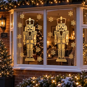 May include: Two gold-colored nutcracker decals and multiple snowflake decals applied to a window pane. The nutcrackers are depicted in traditional military-style uniforms with tall hats, and the snowflakes vary in size and intricate design.