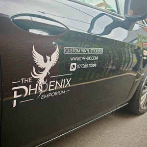 Pode incluir: Porta de carro preta com um logotipo de f&eacute;nix branco e o texto "The Phoenix Emporium". O texto adicional diz "Custom Vinyl Stickers", um endere&ccedil;o de site e um n&uacute;mero de telefone.