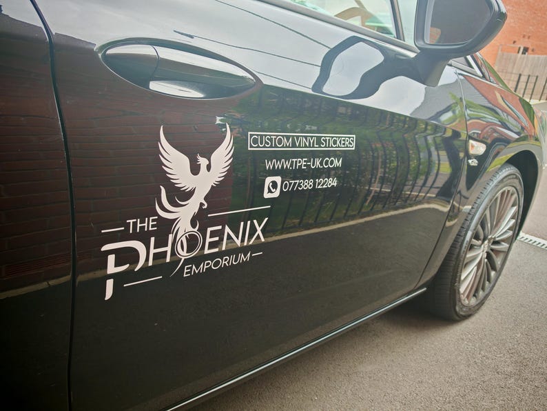 Pode incluir: Porta de carro preta com um logotipo de f&eacute;nix branco e o texto "The Phoenix Emporium". O texto adicional diz "Custom Vinyl Stickers", um endere&ccedil;o de site e um n&uacute;mero de telefone.