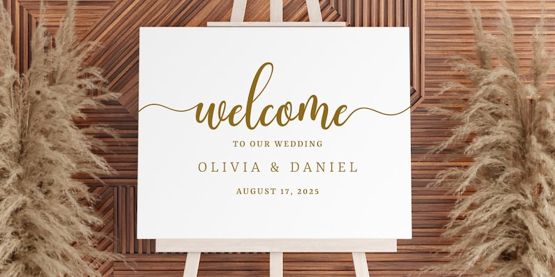 Puede incluir: Un cartel de bienvenida blanco con letras doradas. El cartel dice "Bienvenido a nuestra boda Olivia & Daniel 17 de agosto de 2023".