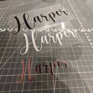 May include: Three vinyl decals with the name "Harper" written in different styles. The top decal is black, the middle decal is white, and the bottom decal is rose gold.