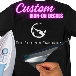 May include: A black t-shirt with a white bird logo and the text "The Phoenix Emporium" and "Custom Iron-On Decals" in pink. A clear sheet of iron-on decal material is being applied to the shirt with an iron.