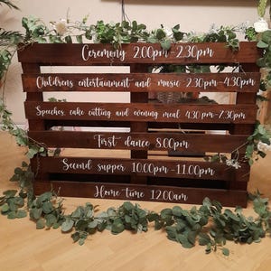 Op de afbeelding: Een houten palletbord met witte letters waarop het schema voor een huwelijksreceptie staat. Het bord bevat de tijden voor de ceremonie, kinderentertainment, speeches, taart snijden, diner, eerste dans en tweede diner. Het bord bevat ook de tijd waarop de gasten naar huis moeten gaan.