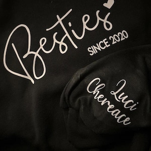 May include: Black t-shirt with white text that says "Besties since 2020" with a heart symbol. A black beanie with white text that says "Luci Chereance" is also shown.
