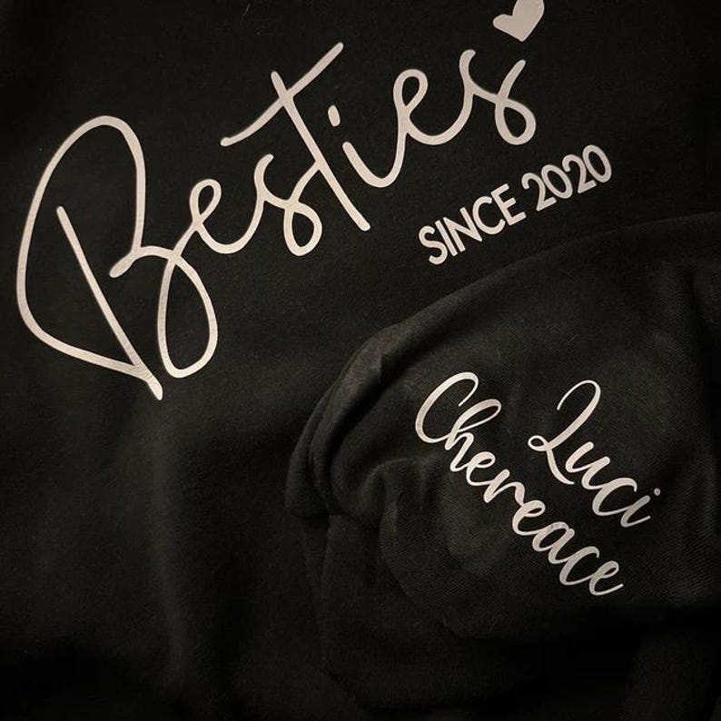May include: Black t-shirt with white text that says "Besties since 2020" with a heart symbol. A black beanie with white text that says "Luci Chereance" is also shown.