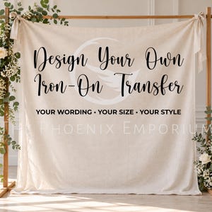 May include: A beige fabric backdrop hanging from a wooden frame with tied corners. The text reads: Design Your Own Iron-On Transfer, YOUR WORDING • YOUR SIZE • YOUR STYLE, and THE PHOENIX EMPORIUM. A faint white phoenix logo is centred behind the text.