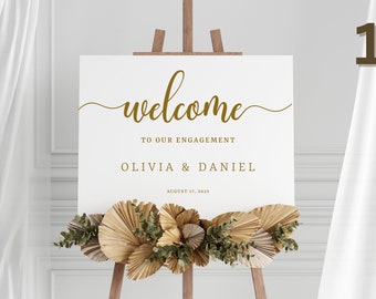 May include: A white welcome sign with gold lettering on an easel. The sign reads "Welcome to our engagement Olivia & Daniel August 17, 2023". The sign is decorated with dried palm leaves and greenery.