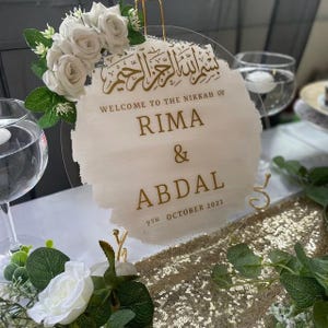 Pu&ograve; includere: Un cartello in acrilico bianco con lettere dorate che annuncia il Nikkah di Rima & Abdal il 7 ottobre 2023. Il cartello &egrave; decorato con fiori bianchi ed &egrave; su un supporto dorato.