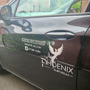 Pode incluir: Uma porta de carro preta com adesivos de vinil personalizados brancos. O texto inclui um site e um n&uacute;mero de telefone. Um logotipo de f&ecirc;nix branco &eacute; exibido abaixo do texto "THE PHOENIX EMPORIUM".