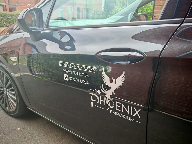 Pode incluir: Uma porta de carro preta com adesivos de vinil personalizados brancos. O texto inclui um site e um n&uacute;mero de telefone. Um logotipo de f&ecirc;nix branco &eacute; exibido abaixo do texto "THE PHOENIX EMPORIUM".