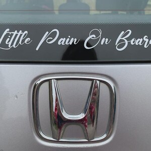 Pode incluir: Um carro prateado com uma faixa preta que diz "Little Pain On Board" em escrita cursiva branca. O emblema da Honda &eacute; vis&iacute;vel abaixo da faixa.