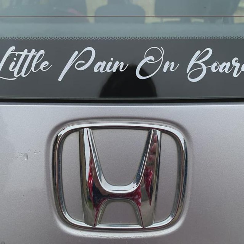Pode incluir: Um carro prateado com uma faixa preta que diz "Little Pain On Board" em escrita cursiva branca. O emblema da Honda &eacute; vis&iacute;vel abaixo da faixa.