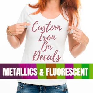 May include: A white t-shirt with the text "Custom Iron On Decals" in pink script. The text is on the front of the shirt, and the person wearing the shirt is pointing to it. The bottom of the image has a banner that says "Metallics & Fluorescent".