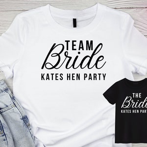 May include: Two white t-shirts with black text. One shirt says "Team Bride Kates Hen Party" and the other says "The Bride Kates Hen Party".