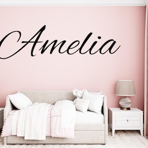 May include: A bedroom scene with a pink wall featuring the name "Amelia" in black script. The room includes a bed with white and pink bedding, two white bedside tables with lamps, and a patterned rug.