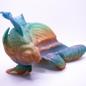 May include: A colorful, stylized fish figurine in shades of blue, green, orange, and yellow. The sculpture has detailed fins and a textured body, with a gradient effect across its form. The fish is positioned on a white surface.