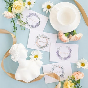 May include: Four white thank you cards with floral wreaths and the text "thank you" in gold lettering. The cards are surrounded by white and pink flowers, a white ceramic bust, and a gold ribbon on a light blue background.