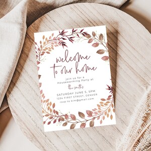 May include: A white invitation card with the text "welcome to our home" in a handwritten font, surrounded by a floral wreath in shades of brown and burgundy. The card includes details for a housewarming party.