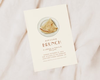 May include: A beige invitation card with brown text announcing a brunch to celebrate a bride-to-be named Olivia June. The invitation includes the date, time, and location of the event. A watercolour illustration of a crepe on a plate is featured at the top of the card.