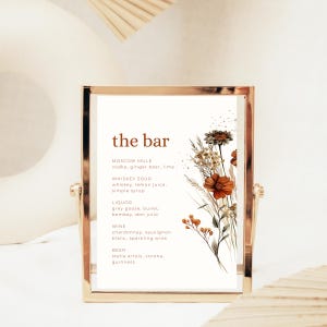 以下が含まれることがあります： 「the bar」と茶色の文字が書かれた金色のフレームの看板。モスコミュール、ウイスキーサワー、さまざまなリキュール、ワイン、ビールなど、カクテルと飲み物のオプションがリストされています。茶色とオレンジ色のフローラルの水彩イラストが右側を飾っています。