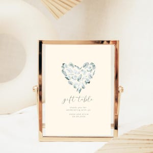May include: A gold-framed sign with a light blue watercolor heart design. The sign reads "gift table" and includes the text "thank you for celebrating with us steve and olivia 05.05.2023".